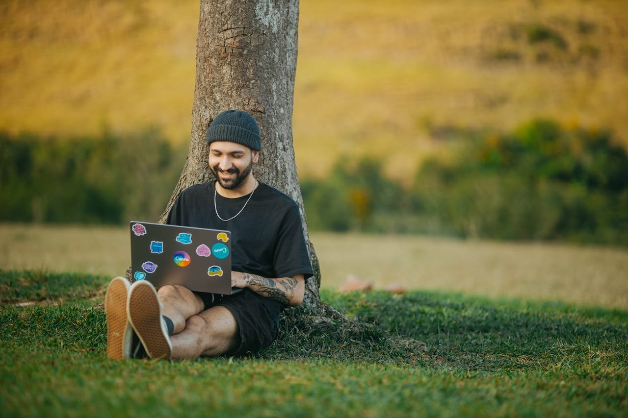 A cheerful man sitting under a tree working on a laptop in a serene rural setting.