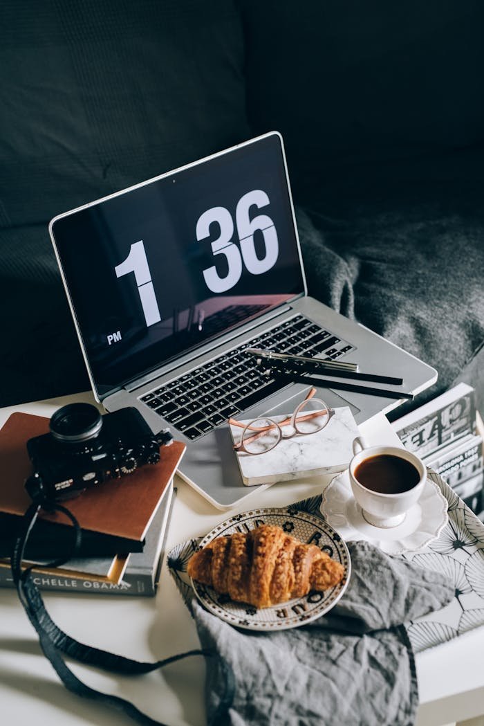 gallery-6 A cozy home office setup with a laptop, coffee, croissant, and books on a sofa.