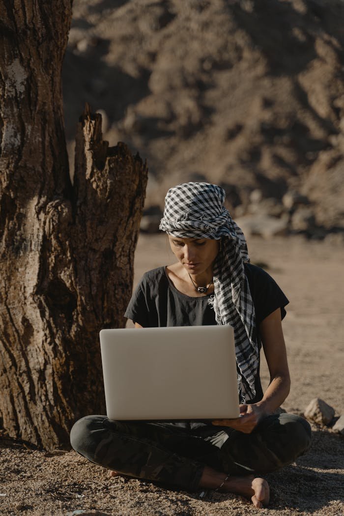 hero-img-02 Young woman sitting under tree working remotely on laptop in arid landscape.