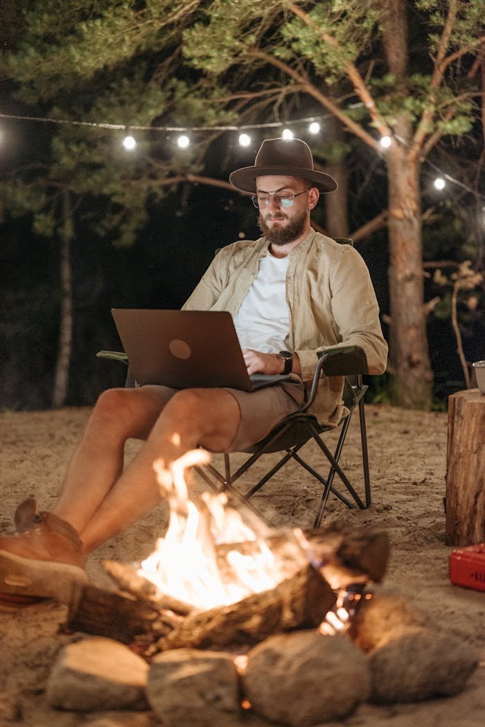 Bearded man working on a laptop in the forest, enjoying the campfire's warmth and solitude.